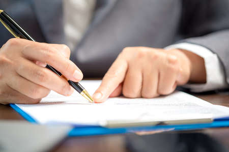 Workplace closeup person professional businesswoman sitting at desk hold pen signing or signature contract paper. Employee woman writing agreement document on paperwork form corporate at work officeの写真素材