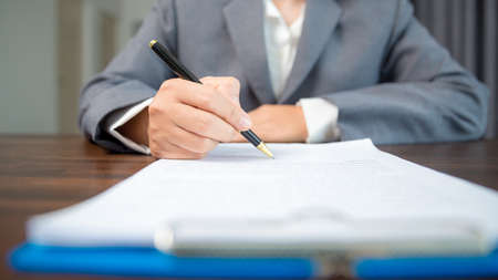 Workplace closeup person professional businesswoman sitting at desk hold pen signing or signature contract paper. Employee woman writing agreement document on paperwork form corporate at work officeの写真素材