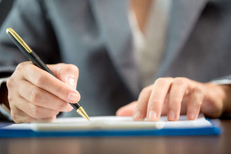 Workplace closeup person professional businesswoman sitting at desk hold pen signing or signature contract paper. Employee woman writing agreement document on paperwork form corporate at work officeの写真素材