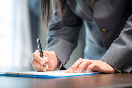 Workplace closeup person professional businesswoman sitting at desk hold pen signing or signature contract paper. Employee woman writing agreement document on paperwork form corporate at work officeの写真素材