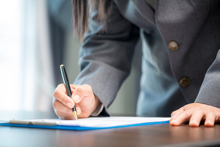 Workplace closeup person professional businesswoman sitting at desk hold pen signing or signature contract paper. Employee woman writing agreement document on paperwork form corporate at work officeの写真素材