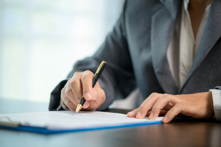 Workplace closeup person professional businesswoman sitting at desk hold pen signing or signature contract paper. Employee woman writing agreement document on paperwork form corporate at work officeの写真素材