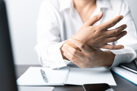 A business office worker is an asian woman is sitting in front of the laptop computer. A businesswoman stressing her body part fingers, hands arm. A femaleOffice manager is exhausted at her workplace.の写真素材