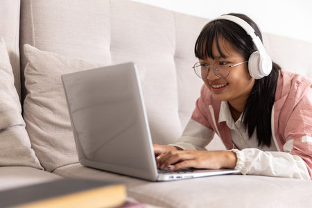 Focused Asian teenage girl wearing headphones writing study notes with laptop and book, serious high...
