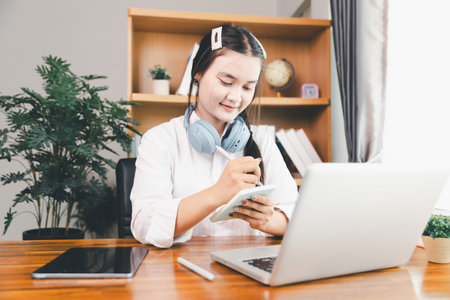 Asian girl student wear headphones learning online watching webinar class looking at laptop computer elearning remote lesson making notes or video calling virtual conference meeting teacher at home.の写真素材