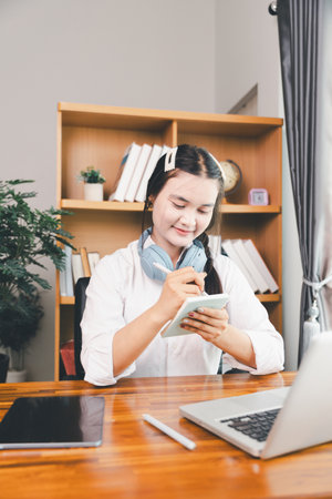 Asian girl student wear headphones learning online watching webinar class looking at laptop computer elearning remote lesson making notes or video calling virtual conference meeting teacher at home.の写真素材
