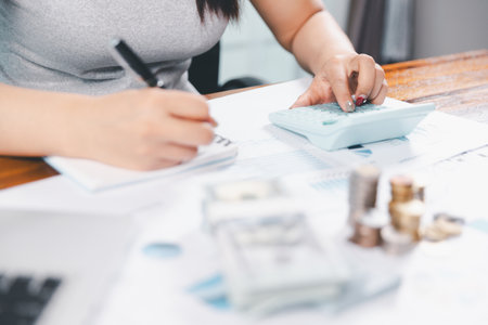 Young Asian woman making stack of coin. invest save finance concept, saving money, investment. dollar bills on table with pile of coins and banks calculator, money to save and invest to make income.の写真素材
