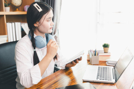 Asian girl student wear headphones learning online watching webinar class looking at laptop computer elearning remote lesson making notes or video calling virtual conference meeting teacher at home.の写真素材