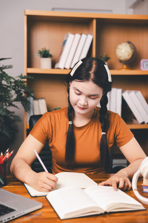 Asian girl doing homework and studying at home. woman using laptop at desk write notes watching webinar, study online, look at learn web class or having virtual call meeting remote working from home.の写真素材