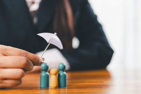 Hands protecting family wooden model for insurance and assurance life concept. Businessman hand gesture protecting wooden domino falling to family dolls for insurance and assurance.の写真素材