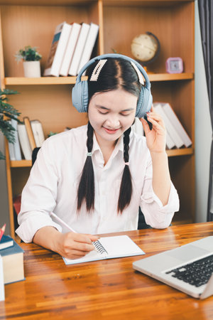 Asian girl student wear headphones learning online watching webinar class looking at laptop computer elearning remote lesson making notes or video calling virtual conference meeting teacher at home.の写真素材