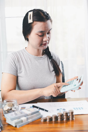 Young Asian woman making stack of coin. invest save finance concept, saving money, investment. dollar bills on table with pile of coins and banks calculator, money to save and invest to make income.の写真素材