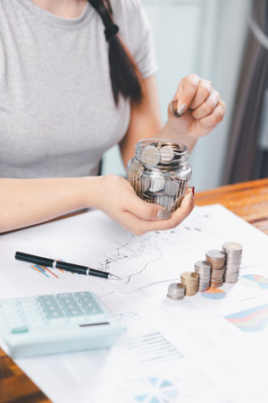 Young Asian woman making stack of coin. invest save finance concept, saving money, investment. dollar bills on table with pile of coins and banks calculator, money to save and invest to make income.の写真素材