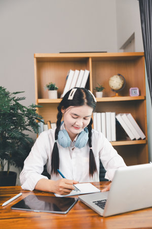 Asian girl student wear headphones learning online watching webinar class looking at laptop computer elearning remote lesson making notes or video calling virtual conference meeting teacher at home.の写真素材