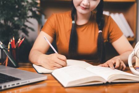 Asian girl doing homework and studying at home. woman using laptop at desk write notes watching webinar, study online, look at learn web class or having virtual call meeting remote working from home.の写真素材