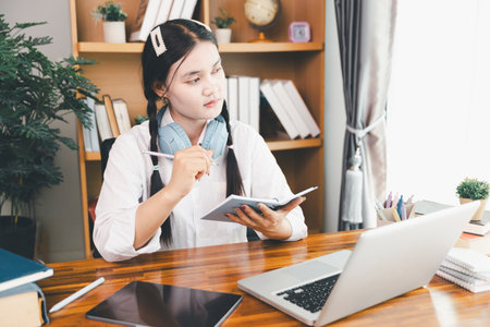 Asian girl student wear headphones learning online watching webinar class looking at laptop computer elearning remote lesson making notes or video calling virtual conference meeting teacher at home.の写真素材