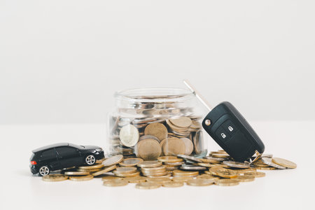 Car key fob and toy car beside a coin jar and scattered change on white background, symbolizing saving for a vehicle, down payment budgeting, auto loan planning and smart finance goals.の写真素材