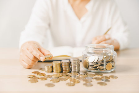 Close-up of hand putting coin into clear glass jar filled with change on bright desk. Minimal home...