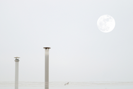 Two smokestacks and the moon while a seagull is flying crossing a grey sky. Empty copy space for editor's text.の写真素材