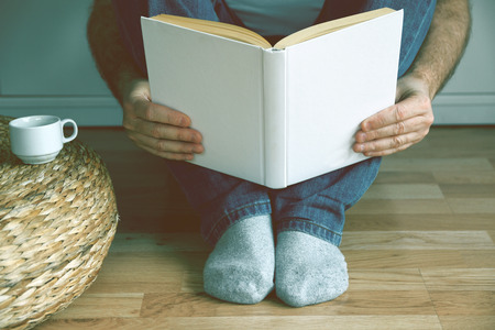 Young man sitting on the wooden floor drinking coffee while reading a book. Empty copy space for...