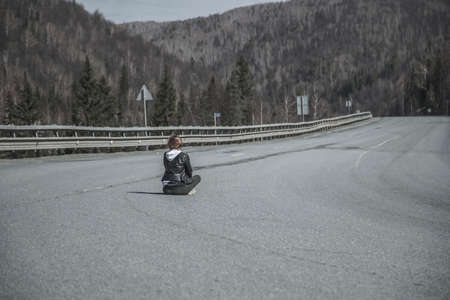 A beautiful girl is sitting on an empty road. Mountains and sky in the distance. Road without cars. Girl in the middle of the road.の写真素材