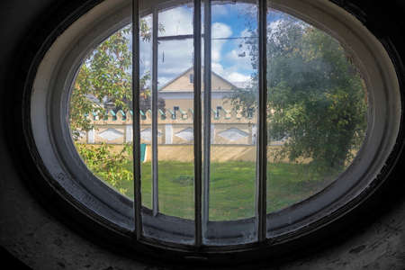 View of the trees and the old building from the window of an abandoned hospital. Round window. Beautiful nature outside the window.の写真素材