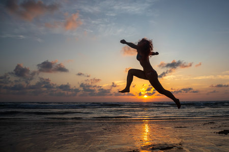 Silhouette of young woman jumping on the beach at sunset.の写真素材