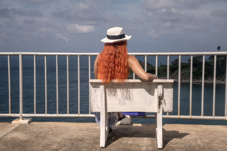 Red-haired girl in a white hat sits on a bench by the sea.の写真素材