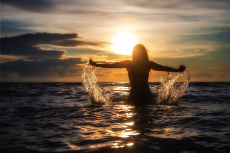 Silhouette of a girl in the water splashes with her hands on the sunset backgroundの写真素材