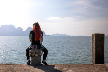 A young girl with long red hair sits on a concrete pier and looks at the sea.の写真素材