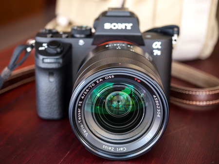 BANGKOK - FEBRUARY 19: Sony digital camera model A7II with normal Carl Zeiss zoom lens 24-70 mm f4 in natural light on red wooden table, focus on front surface of the lens, was taken on February 19, 2015, in Bangkok Thailand.のeditorial素材