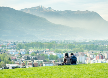 Two unrecognized people sit together on green grass with Bellinzona cityscape view and surrounding mountains in Switzerland, under cloudy blue skyの写真素材