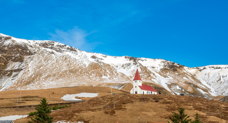 Small church with red roof in Vik town at top of small hill with snow mountains in background in winter season in Icelandのeditorial素材