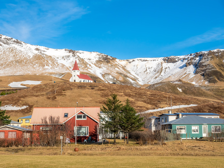 House in village and small church at top of small hill near black sand beach in Vik town in Iceland with snow mountains in background under clear blue sky in winter seasonの写真素材