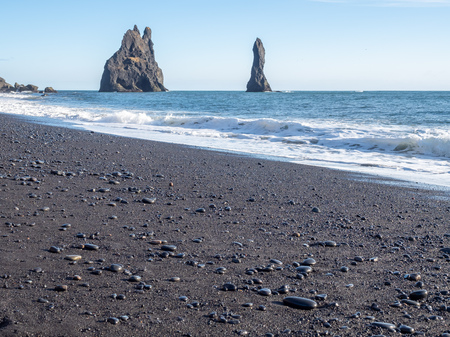 Reynisfjara black sand beach is landmark of Vik town in Iceland, fine black sand with small stones on beach with mountain and islands in background, under blue sky in winter seasonの写真素材