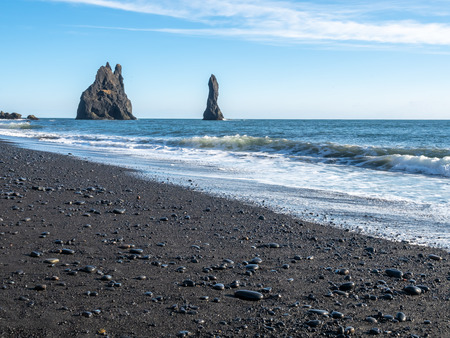 Reynisfjara black sand beach is landmark of Vik town in Iceland, fine black sand with small stones on beach with mountain and islands in background, under blue sky in winter seasonの写真素材