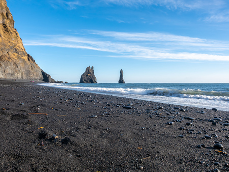 Reynisfjara black sand beach is landmark of Vik town in Iceland, fine black sand with small stones on beach with mountain and islands in background, under blue sky in winter seasonの写真素材