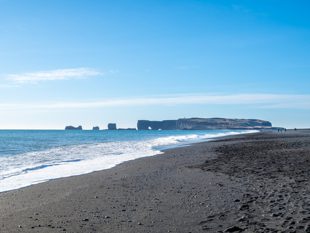 Reynisfjara black sand beach is landmark of Vik town in Iceland, fine black sand with small stones on beach with mountain and islands in background, under blue sky in winter seasonの写真素材