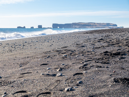 Reynisfjara black sand beach is landmark of Vik town in Iceland, fine black sand with small stones on beach with mountain and islands in background, under blue sky in winter seasonの写真素材