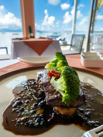 Sirloin beef steak with red wine sauce served with potato and broccoli on white round dish in European restaurant, with sea view in backgroundの写真素材