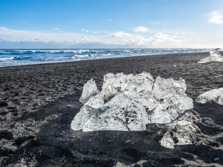Ice pieces, glitter like diamonds, on black sand beach in winter season in Iceland, under cloudy blue skyの写真素材