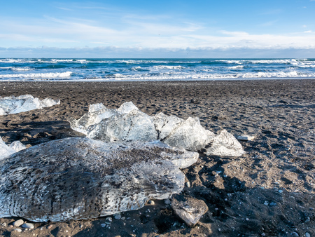Ice pieces, glitter like diamonds, on black sand beach in winter season in Iceland, under cloudy blue skyの写真素材