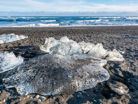 Ice pieces, glitter like diamonds, on black sand beach in winter season in Iceland, under cloudy blue skyの写真素材