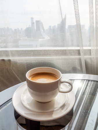 Cup of coffee on round glass table with natural window light in hotel room with cityscape view behind transparent curtainの写真素材
