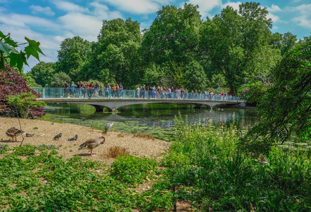 St. James, London, UK - June 8, 2018:  Lots of people crossing over the blue bridge in St. Jame's Park on a bright sunny summer day.  Shows a heron and geese in the foreground.のeditorial素材