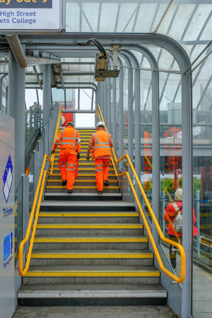 Poplar, London, UK - August 18, 2018: Two workmen dressed in orange hi-viz clothing  and hard hats walking up the stairs at Poplar station.のeditorial素材