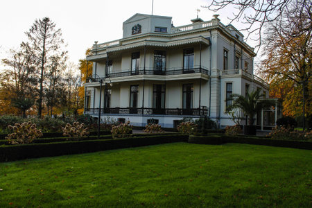 A beautiful white mansion with a classic architectural style, surrounded by lush green lawns and neatly trimmed hedges. The building features balconies and large windows, set against a backdrop of autumn trees.の写真素材