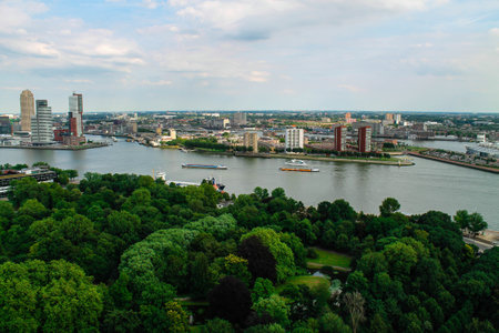 Aerial view of a river winding through a city with modern buildings and lush green parks along the banks. Boats are seen on the water, and the skyline features a mix of architecture under a partly cloudy sky.の写真素材