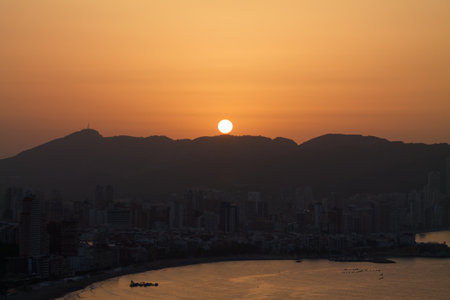 A serene sunset over a coastal city, with the sun setting behind mountains. The skyline features tall buildings and a calm beach, reflecting warm orange hues.の写真素材