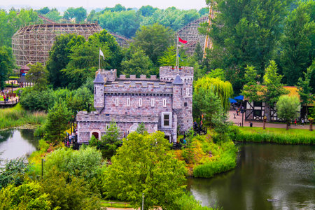 Netherlands, Biddinghuizen - July 15, 2014 - A picturesque view of a small stone castle surrounded by lush greenery and a serene pond. In the background, amusement park rides are visible, adding a playful contrast to the tranquil scene.の写真素材